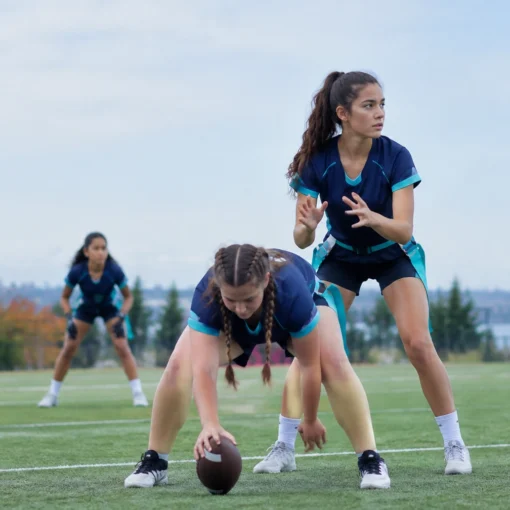 Girl crouches to snap a football on a grassy field while teammates prepare in blue uniforms in the background.