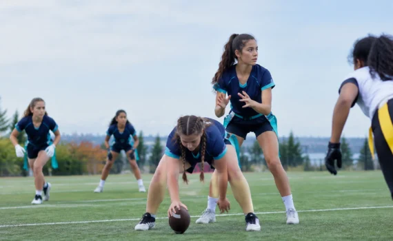 Girl crouches to snap a football on a grassy field while teammates prepare in blue uniforms in the background.